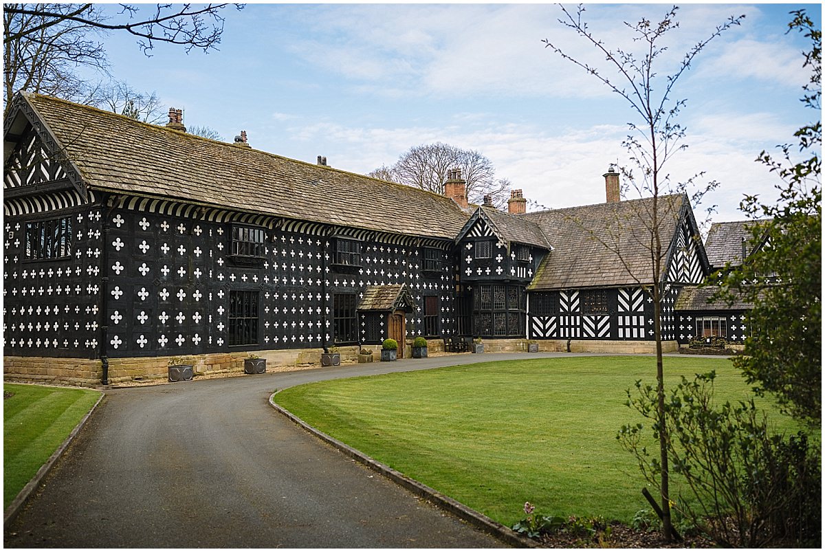 A collection of historic half-timbered buildings with decorative black and white facades, surrounded by a well-manicured lawn and a curved pathway at Samlesbury Hall.
