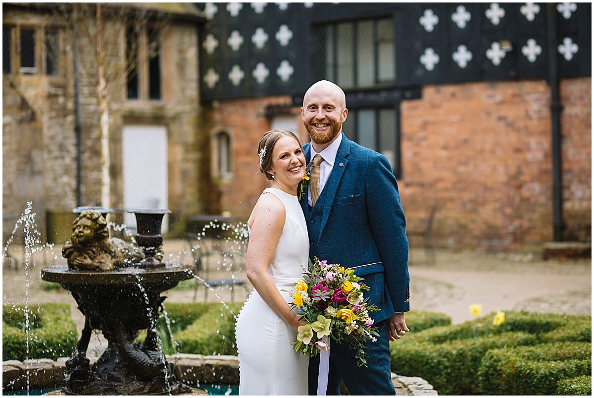 A smiling bride and groom pose for a photo in front of a historic building with black and white details, standing beside a water fountain with a cherub statue. The bride holds a colorful bouquet of flowers.