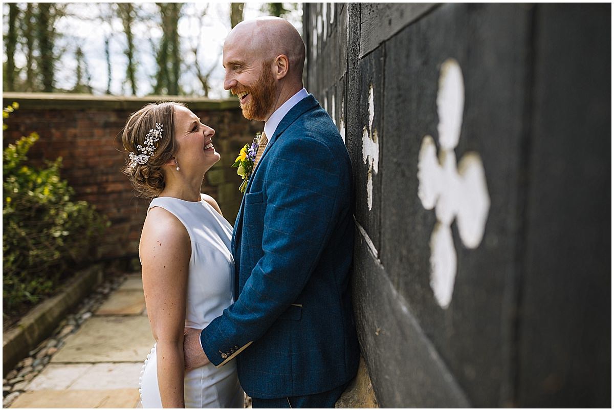 A happy couple on their wedding day at Samlesbury Hall, leaning against a dark wall with peeling paint, sharing a joyful moment. The bride in a white sleeveless dress with a floral hair accessory smiles at the groom in a blue suit, who is smiling back at her.
